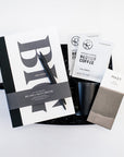 Wit and Delight Big Idea Box, a black Miir camp mug with a lid, two black pens, two packets of Reborn Pour Over Colombia Coffee, and a large Mast dark chocolate bar arranged in a black gifting box with black paper shred.
