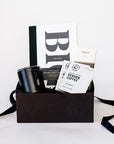 A black box with a Wit and Delight Big Idea Box, a black Miir camp mug with a lid, two black pens, two packets of Reborn Pour Over Colombia Coffee, and a large Mast dark chocolate bar.