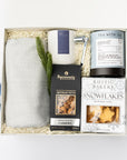 top down photo of a holiday themed cream gift box featuring a light gray herringbone throw blanket, a white ceramic candle with blue label named Silent Night, metal silver candle snuffer, grey canister of peppermint tea sachets, black with silver foil detail box of artisanal cashews, white box of shortbread cookies snowflake shaped with silver foil snowflakes print, preserved spruce leaf ornament. All items are nicely nested on ivory thin shredded paper with winter-blue ribbon around the box.