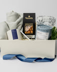 top down photo of a holiday themed cream gift box featuring a light gray herringbone throw blanket, a white ceramic candle with blue label named Silent Night, metal silver candle snuffer, grey canister of peppermint tea sachets, black with silver foil detail box of artisanal cashews, white box of shortbread cookies snowflake shaped with silver foil snowflakes print, preserved spruce leaf ornament. All items are standing, staged inside the box with winter-blue ribbon around the box.