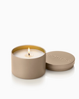 A lit candle in a matte taupe metal tin with its lid resting beside it on a clean white background. The embossed brand name 'APOTHEKE' is visible on the lid.