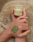 Hand holding a jar of Balancing Face Mask with green mask on wrist against a stone background