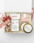 The Be My Bridesmaid Gift Box is featured in our signature white gift box filled with ivory shred. From left to right, it includes a dried floral bundle, a metallic rose gold “XOXO” matchbox in the center, “I Couldn’t Tie the Knot Without You” earrings, champagne bubbly gummy bears, and a “Will You Be My Bridesmaid?” candle in a gold tin. Displayed on a white background with a light pink ribbon surrounding it.