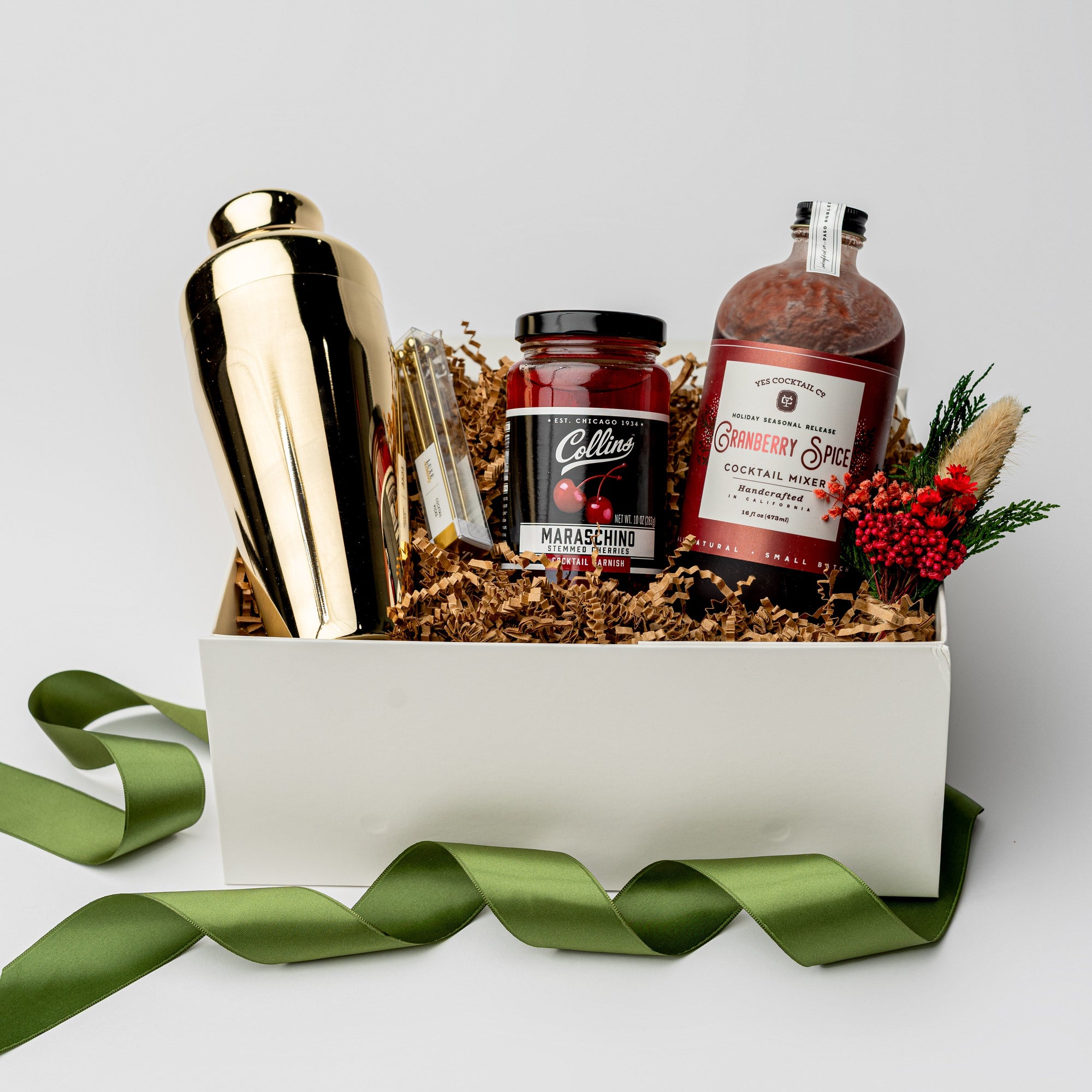 Berry Merry holiday gift box, in a white box with brown shred and a green ribbon around it. Inside of the box in the left gold cocktail shaker, in the center set of four gold cocktail picks, stemmed maraschino cherries cocktail garnish , cranberry spice cocktail mixer red and white label , and on the left mini floral bundle on a white background.
