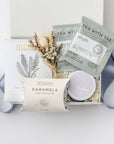 The Better Days Ahead sympathy Gift Box is featured in our signature white gift box filled with ivory shred. From left to right, it includes a Formulary Eucalyptus & Peppermint mineral soak, Cape Cod sea salt caramels, a dried floral bundle in the center, and on the right, two calming tea sachets and a “Thinking of You” candle. Displayed on a white background with a light blue ribbon surrounding it.