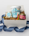 Front view of a white box birthday gift box with brown shreds and a blue ribbon, featuring vanilla ice cream mix, caramel sauce, confetti caramels, a gold spoon, and four mini birthday candles in red, blue, green, and yellow.