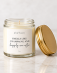 A 4 oz glass jar candle with cream-colored wax and a white label reading 'fkblossom SMELLS LIKE CHAMPAGNE AND happily ever after' in black text. The gold metal lid is placed beside the open jar on a light marble surface.