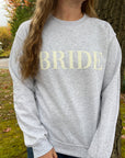 Gray crewneck sweatshirt with large white-embroidered “BRIDE” lettering across the chest worn by a young woman with long hair on a nature background.