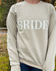 Nude crewneck sweatshirt with large white-embroidered “BRIDE” lettering across the chest worn by a young woman with long hair on a nature.