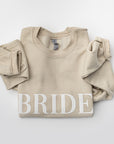 Folded nude sweatshirt with “BRIDE” embroidered in white thread across the chest, displayed on a white background.