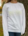 White crewneck sweatshirt with large white-embroidered “BRIDE” lettering across the chest worn by a young woman with long hair on a nature.