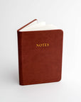 Burgundy pocket softcover notebook featured on a white background. With Notes in gold in front of it.
