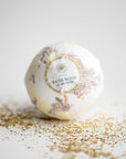 Chamomile bath bomb ,round, in its transparent covering and label that has a chamomile flower and black lettering. Surrounded by chamomile pieces.