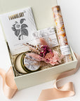 A front view of the Coming Up Roses Spa Gift Box featuring, from left to right, a 4 oz. hand-poured Santal & Vanilla candle, a white matchsticks jar, a Formulary 55 Rosehip & Clay facial mask, a white tea shea butter guest soap, rose bath soaking salts, and a handmade dried floral mini bouquet. Arranged in a white gift box filled with ivory shred and wrapped with a soft pink ribbon around the box on a white background.