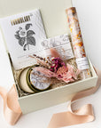 A front view of the Coming Up Roses Spa Gift Box featuring, from left to right, a 4 oz. hand-poured Santal & Vanilla candle, a white matchsticks jar, a Formulary 55 Rosehip & Clay facial mask, a white tea shea butter guest soap, rose bath soaking salts, and a handmade dried floral mini bouquet. Arranged in a white gift box filled with ivory shred and wrapped with a soft pink ribbon around the box on a white background.