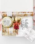 A top view of the Coming Up Roses Spa Gift Box featuring, a 4 oz. hand-poured Santal & Vanilla candle, a white matchsticks jar, a Formulary 55 Rosehip & Clay facial mask, a white tea shea butter guest soap, rose bath soaking salts, and a handmade dried floral mini bouquet. Arranged in a white gift box filled with ivory shred and wrapped with a soft pink ribbon around the box on a white background.