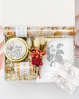A top view of the Coming Up Roses Spa Gift Box featuring, a 4 oz. hand-poured Santal & Vanilla candle, a white matchsticks jar, a Formulary 55 Rosehip & Clay facial mask, a white tea shea butter guest soap, rose bath soaking salts, and a handmade dried floral mini bouquet. Arranged in a white gift box filled with ivory shred and wrapped with a soft pink ribbon around the box on a white background.