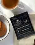 Copper Cow Coffee, Vietnamese Coffee, is featured in its black sachet with white lettering. In the background, a marble-like table with cookies in a bowl, a clear mug with milk and coffee, a napkin, and a rose gold spoon.