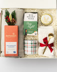 Cozy at home holiday gift box is featured  in a white box with ivory shred on the back with red ribbon underneath it.  On the left gray throw blanket with a moonstruck nutty milk chocolate cinnamon chocolate bar, and mini dried floral bundle. In the middle cozy night herbal tea, plaid mug green and red stripes. On the right spruce gold candle, white cloud socks tied in red ribbon and gold platted stirring spoon. On a white background.