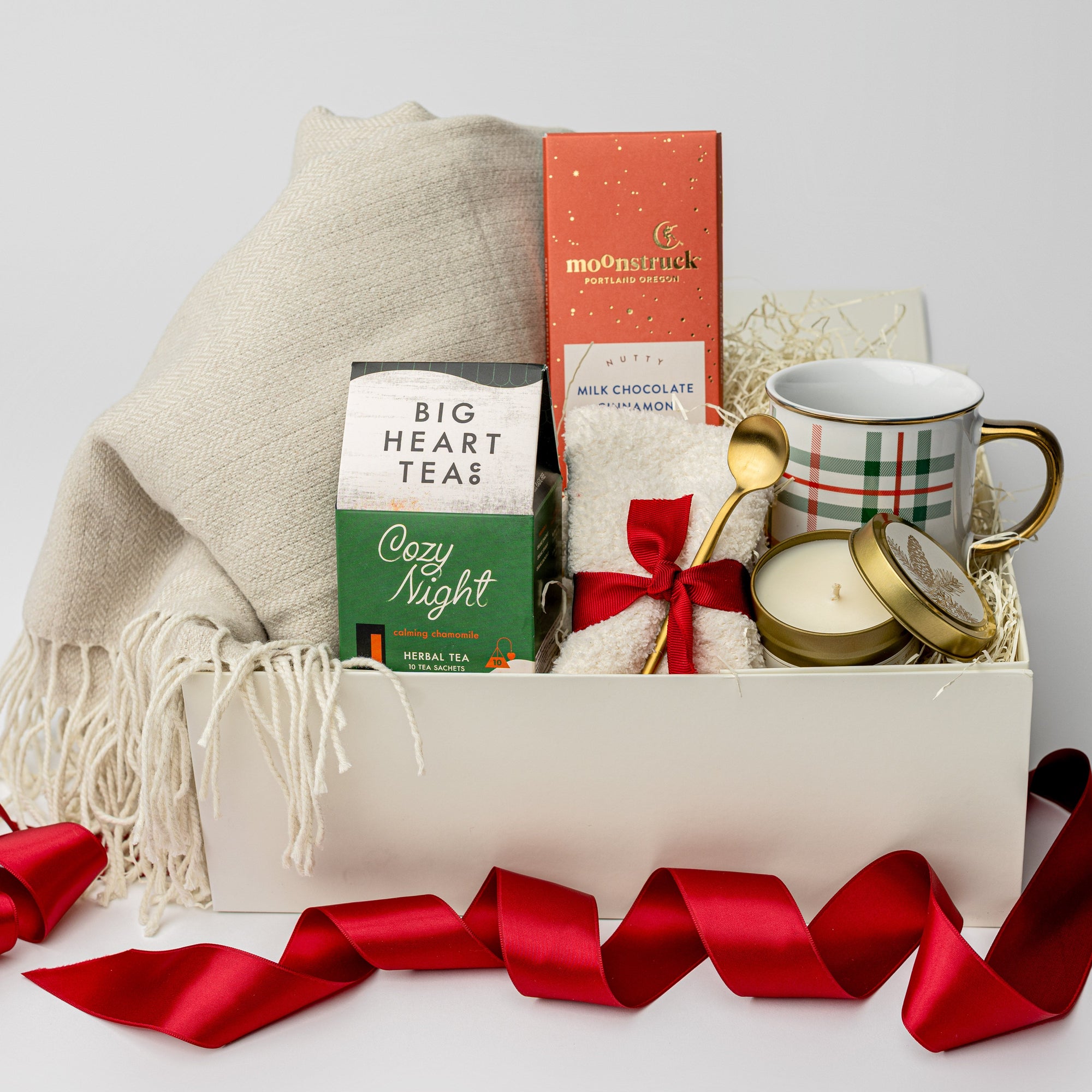 Cozy at Home Holiday Gift Box is featured in a white box wrapped with a red ribbon. Inside the box, on the left, there is a throw blanket and a Cozy Night herbal tea. In the middle, a Moonstruck Nutty Milk Cinnamon Chocolate Bar and a pair of white cozy socks tied with a red ribbon and a gold-plated stirring spoon attached. On the right, a plaid mug with red and green lines and a Spruce gold candle. The image is displayed on a white background.