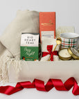 Cozy at Home Holiday Gift Box is featured in a white box wrapped with a red ribbon. Inside the box, on the left, there is a throw blanket and a Cozy Night herbal tea. In the middle, a Moonstruck Nutty Milk Cinnamon Chocolate Bar and a pair of white cozy socks tied with a red ribbon and a gold-plated stirring spoon attached. On the right, a plaid mug with red and green lines and a Spruce gold candle. The image is displayed on a white background.