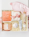 A white gift box filled with pink ribbon and ivory paper shred on a white background. Inside the box are a pink satin sleep mask, Cup of Love rose tea for two, an 11oz hand-poured soy double-wick candle in a keepsake glass jar labeled “Sweet Fig,” plush ivory cloud socks, a tube of shea butter hand crème in the scent Santal Vanille, a rose petal bath bomb, and a jar of rose bath salts — a cozy, self-care “Cup of Love” gift box.