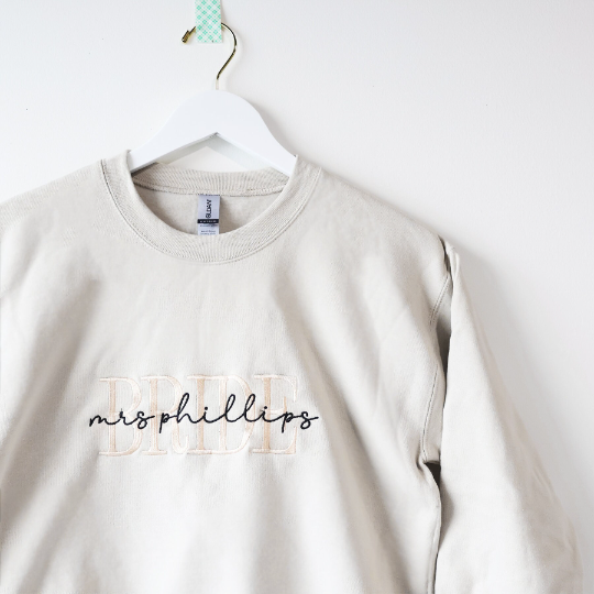Custom embroidered cream-colored crewneck sweatshirt. Embroidered on the front are the words "BRIDE" in a block-letter font in ivory thread and the words "mrs Phillips" in a more delicate cursive style in black thread on a white background, hung on a white hanger.