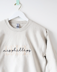 Custom embroidered cream-colored crewneck sweatshirt. Embroidered on the front are the words "BRIDE" in a block-letter font in ivory thread and the words "mrs Phillips" in a more delicate cursive style in black thread on a white background, hung on a white hanger.