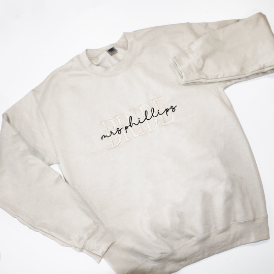 Custom embroidered cream-colored crewneck sweatshirt. Embroidered on the front are the words "BRIDE" in a block-letter font in ivory thread and the words "mrs Phillips" in a more delicate cursive style in black thread on a white background.