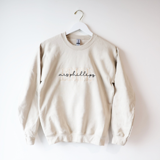 A cream-colored crewneck sweatshirt. Embroidered on the front are the words "BRIDE" in a block-letter font in ivory thread and the words "mrs Phillips" in a more delicate cursive style in black thread on a white background, hung on a white wooden hanger.