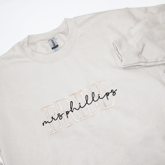 Close up custom embroided  cream-colored crewneck sweatshirt. Embroidered on the front are the word "BRIDE" in a block-letter font in ivory thread and the words "mrs. Phillips" in a more delicate cursive style in black thread on a white background.