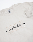 Close up custom embroided cream-colored crewneck sweatshirt. Embroidered on the front are the word "BRIDE" in a block-letter font in ivory thread and the words "mrs. Phillips" in a more delicate cursive style in black thread on a white background.