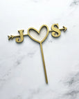 Custom initial wooden cake topper is featured with the initials J heart S with an arrow across in gold color in a marble like background.