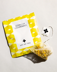 Egyptian chamomile tea sachet is featured in its square shape sachet with yellow hexagons white label with black lettering and in front of the sachet the tea itself out of its package on a white background.