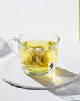 Egyptian chamomile tea sachet featured in a clear glass mug showcasing the tea bag inside that gives the water a warm yellow color on a white background.