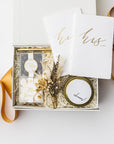 Engagement celebration gift box displayed on a white background with ivory shred and an orange ribbon, containing a set of foil-stamped vow books, two gold minimalist bullet pens, Mrs. champagne bubbles gummies, Mr. cordials caramels, a 4oz hand-poured “Congrats” candle, and a dried floral mini bouquet.