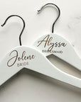 Engraved bride hangers are featured, two white hangers with silver hooks personalized Jolene -Bride and Alyssa - Bridesmaid on a marble-like background.