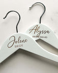 Engraved bride hangers are featured, two white hangers with silver hooks personalized Jolene -Bride and Alyssa - Bridesmaid on a marble-like background.