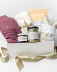 A front view of a postpartum care gift set featuring a cotton nursing gown, Dandelion Tallow Balm, Wildcraft postpartum tea, Wildcraft Recover sitz bath, cozy socks, and a heat pad, all arranged in a white gift box with ivory shred and accented with a green ribbon on a white background.