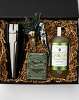 Fireside Spirits Gift Box is featured with a black box and a green ribbon underneath. Inside the box, on the left, there is a gunmetal-plated cocktail shaker. In the middle, a floral bundle, a gunmetal-plated corkscrew, and Monarch luxury playing cards with green and gold details. On the right, a Ginger Snap cocktail mixer with a white label and green details. All items are arranged in front of brown shred in the back, displayed on a white background.