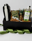 Fireside Spirits Gift Box is featured with a black box and a green ribbon around it. Inside the box, on the left, there is a gunmetal-plated cocktail shaker. In the middle, a floral bundle, a gunmetal-plated corkscrew, and Monarch luxury playing cards with green and gold details. On the right, a Ginger Snap cocktail mixer with a white label and green details. All items are arranged in front of brown shred in the back, displayed on a white background.