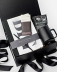 A top view of the Seize the Day Gift Box presented in a black gift box filled with brown shred and tied with a black ribbon, featuring a Big Idea sketchbook, a minimalist black bullet pen, a 12 oz. black vacuum insulated camp mug, a Copper Cow single-serve pour-over coffee, and a MAST organic dark chocolate bar (80% cacao), all arranged on a white background.