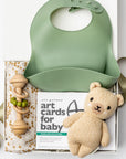 A top view of a Baby Boy Gift Box featuring a knitted cute honey bear, a honeycomb-patterned swaddle, a wooden rattle, woodland animal cards, and a green silicone bib with an engraved name, all arranged in a white gift box with ivory shred and tied with a soft green ribbon on a white background.