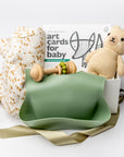A front view of a Baby Boy Gift Box featuring a knitted cute honey bear, a honeycomb-patterned swaddle, a wooden rattle, woodland animal cards, and a green silicone bib with an engraved name, all arranged in a white gift box with ivory shred and tied with a soft green ribbon on a white background.
