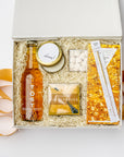 A top view of the Celebration Gift Box featuring a bottle of TÖST Sparkling White Tea, Champagne Bubble Gummies, a Champagne Dark Chocolate Bar, a gold wish sparkler wand, a 4 oz. hand-poured “Cheers” candle, and a deck of Celebrate Conversation Starter Cards, all arranged in a white gift box with ivory shred on a light background and orange ribbon underneath and surrounding the box.