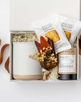 A top view of the Chai Tea Time Gift Box featuring a 12 oz. speckled ceramic mug on the left, an artisan chai tea mini tube and two toffee shortbread cookies in the center, and a handmade dried floral mini bouquet on the right. Arranged in a white gift box with ivory shred and accented with a toffee ribbon crossing underneath the box on a white background.