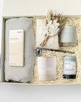 Home Still gift box featuring an 8 oz. Home Sweet Home cedarwood and vanilla candle, silver candle snuffer, peppermint tea mini tube, cloud socks, speckled stoneware cup, herringbone throw blanket, almond and sea salt chocolate, and a handmade dried floral bouquet, beautifully arranged in gift packaging on a white background.