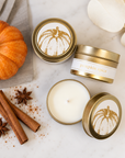 Three gold tin candles with white labels featuring a pumpkin illustration are arranged on a white marble surface with autumn props including a small orange pumpkin, cinnamon sticks, star anise, and cream petals. One candle is open, showing white wax, while the others are closed.