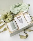 A top view of a soothing spa-themed gift box presented in a white gift box filled with ivory shred and tied with a green ribbon, featuring detox bath salts, eucalyptus & peppermint sea mineral foot soak, a 12 oz. hand-poured eucalyptus & lavender candle in a matte white ceramic jar, a Tillandsia Xerographica live air plant, and a sage silk sleep mask, all arranged on a white background.