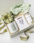 A top view of a soothing spa-themed gift box presented in a white gift box filled with ivory shred and tied with a green ribbon, featuring detox bath salts, eucalyptus & peppermint sea mineral foot soak, a 12 oz. hand-poured eucalyptus & lavender candle in a matte white ceramic jar, a Tillandsia Xerographica live air plant, and a sage silk sleep mask, all arranged on a white background.
